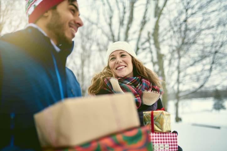 Smiling diverse couple holding Christmas presents while walking through a winter forest